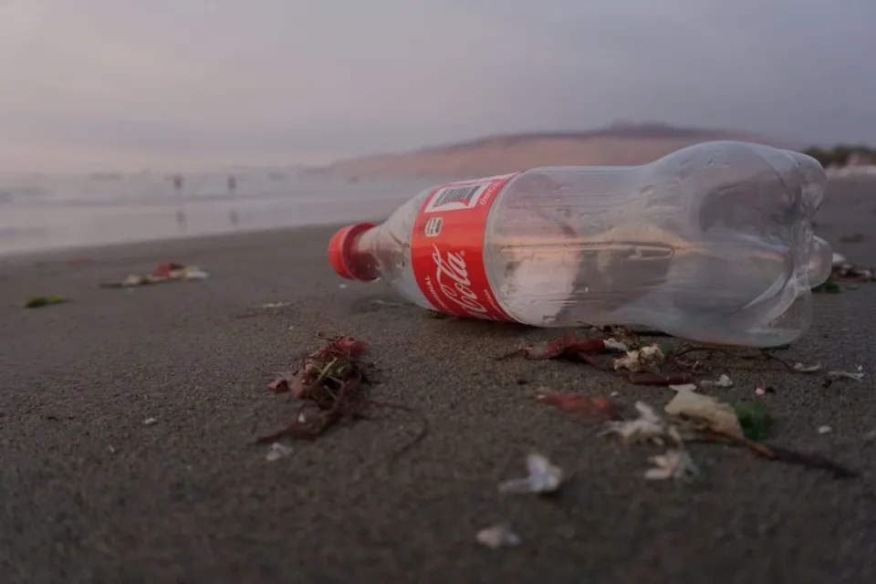 O sticlă de plastic transparentă cu etichetă roșie este întinsă pe nisipul unei plaje sub un cer închis.