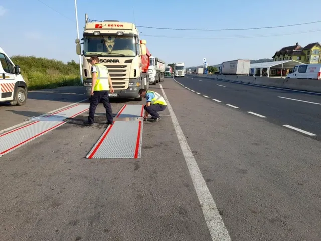 Un bărbat în vestă galbenă stă lângă două rampe metalice de pe asfalt, în timp ce un alt bărbat este aplecat lângă ele.