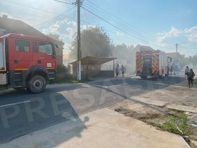 Un camion de pompieri roșu este parcat pe un drum lângă o structură acoperită, în timp ce câțiva oameni stau în apropiere.