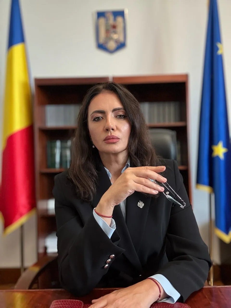 O femeie cu părul lung și întruchipată într-o jachetă neagră stă la o masă în fața unor steaguri și un raft cu cărți.