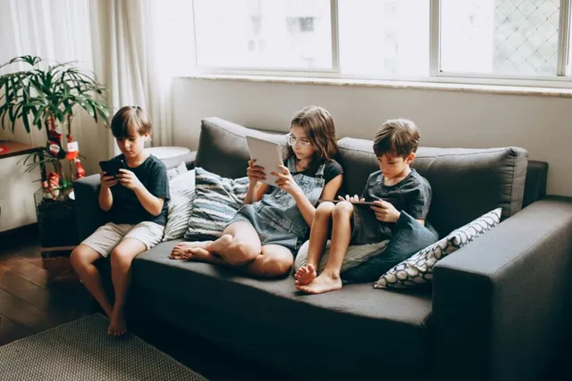 Trei copii stau pe o canapea gri și folosesc dispozitive electronice, precum tablete și telefoane mobile, într-o încăpere luminoasă.