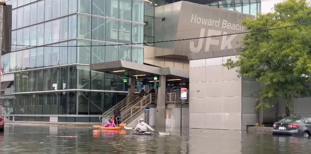 O clădire modernă cu inscriptionatul 'Howard Beach JFK' este înconjurată de ape inundate, în timp ce persoane în canoe navighează prin apă lângă scară.