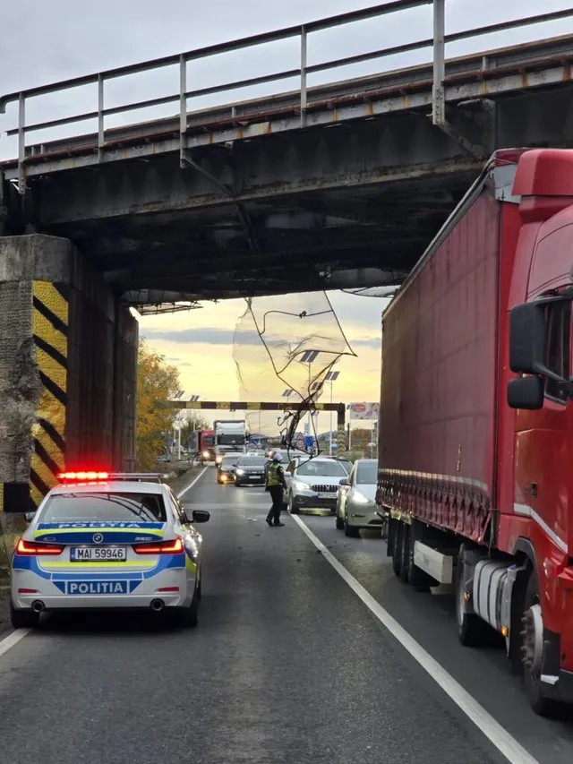 Un vehicul de poliție cu girofaruri aprinse stă pe un drum asfaltat sub un pasaj feroviar, lângă un camion roșu și alte mașini.