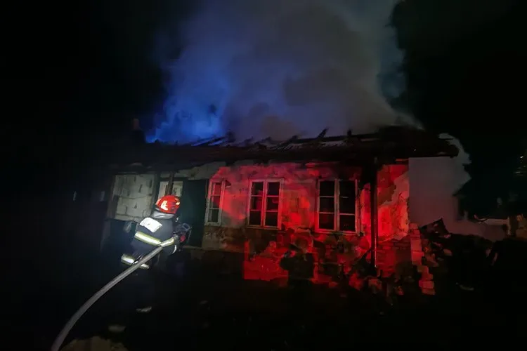 Un pompier cu echipament de protecție și cască roșie stă în fața unei case în flacără, observând fumul dens care se ridică în noapte.
