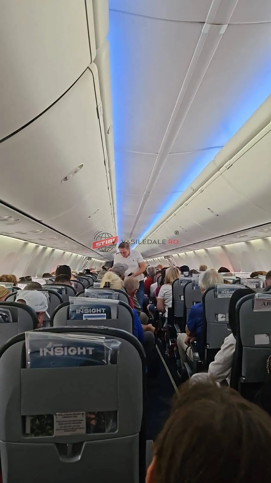 Pasageri sunt așezați în scaunele unui avion, sub un plafon iluminat cu o bandă de lumină albastră.