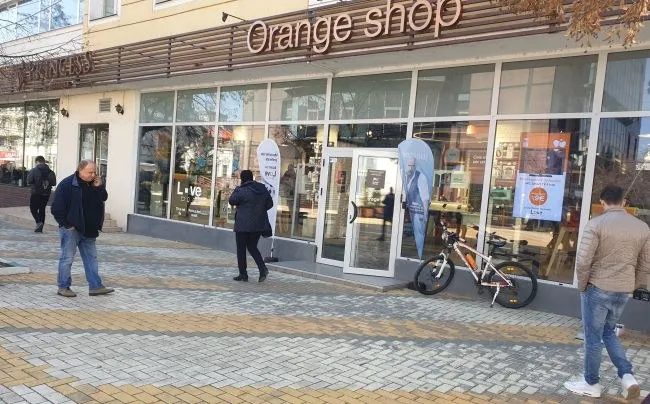 Un bărbat în sacou și pantaloni albaștri merge pe un trotuar de plăci colorate în fața unei vitrine de magazin cu geamuri mari.