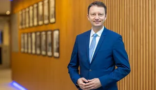 Un bărbat cu părul scurt, purtând un costum albastru și o cămașă albă cu cravată albastră, stă în fața unui perete din lemn cu cadre încadrate.