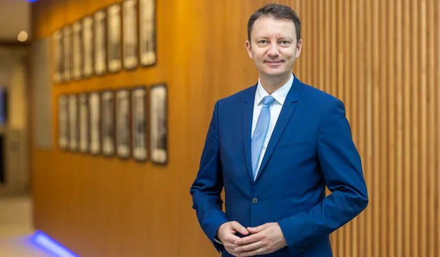 Un bărbat într-un costum albastru și cămașă albă cu cravată deschise la culoare stă în picioare în fața unui perete din lemn cu cadre înrudite.