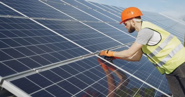 Un bărbat cu cască portocalie și vestă reflectorizantă lucrează la instalarea panourilor solare de pe un acoperiș.