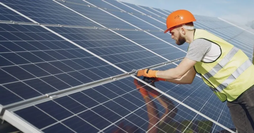 Un bărbat cu cască portocalie și vestă reflectorizantă lucrează la instalarea panourilor solare de pe un acoperiș.