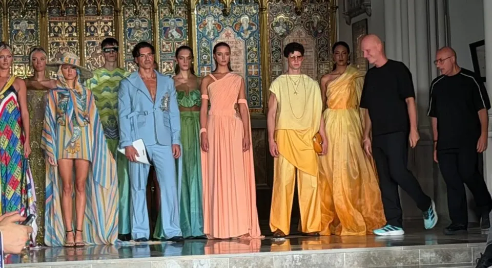 Mai mulți modele poartă diverse haine colorate și voluminoase pe o scenă în fața unui perete decorat cu motive aurii și picturi.
