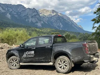 Un pickup negru cu un logo alb pe ușă este parcat pe un teren accidentat în fața unui masiv muntos acoperit de vegetație.