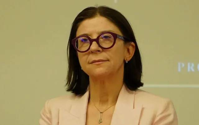 O femeie cu părul scurt și negru poartă ochelari cu rame roșii și un sacou roz deschis.