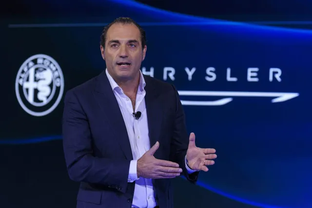 Un bărbat într-un costum închis și cămașă deschisă vorbește cu mâinile ridicate în fața unui fundal albastru cu logourile Alfa Romeo și Chrysler.