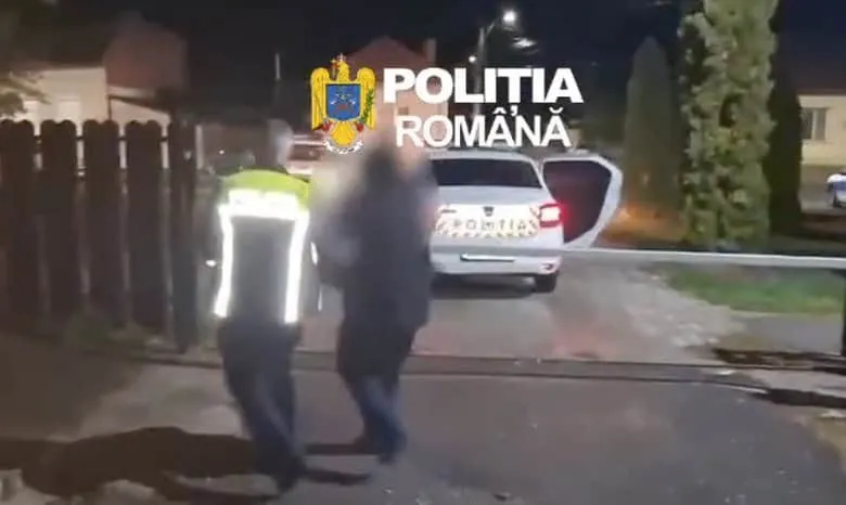 Un agent de poliție în uniformă reflectorizantă stă lângă un vehicul alb cu portierea deschisă pe un drum de noapte.