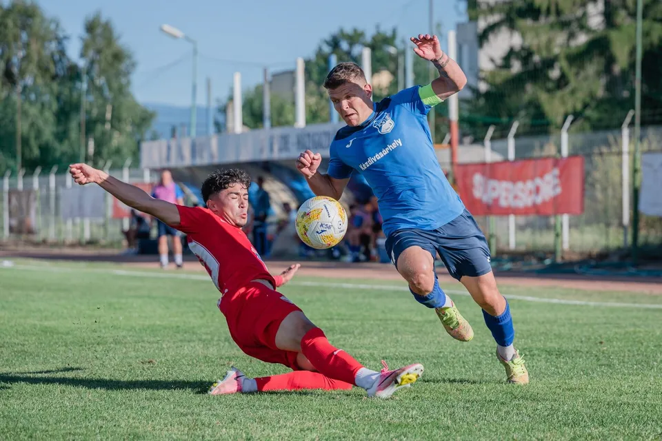 Un jucător în echipament roșu execută o taclă pe terenul de fotbal, în timp ce un adversar în echipament albastru urmărește mingea.