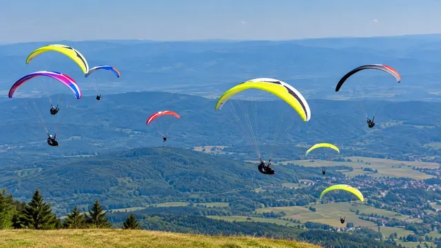 Mai mulți parapantisti cu parapante colorate zboara deasupra unui peisaj montan cu păduri și sate întinse sub un cer senin.