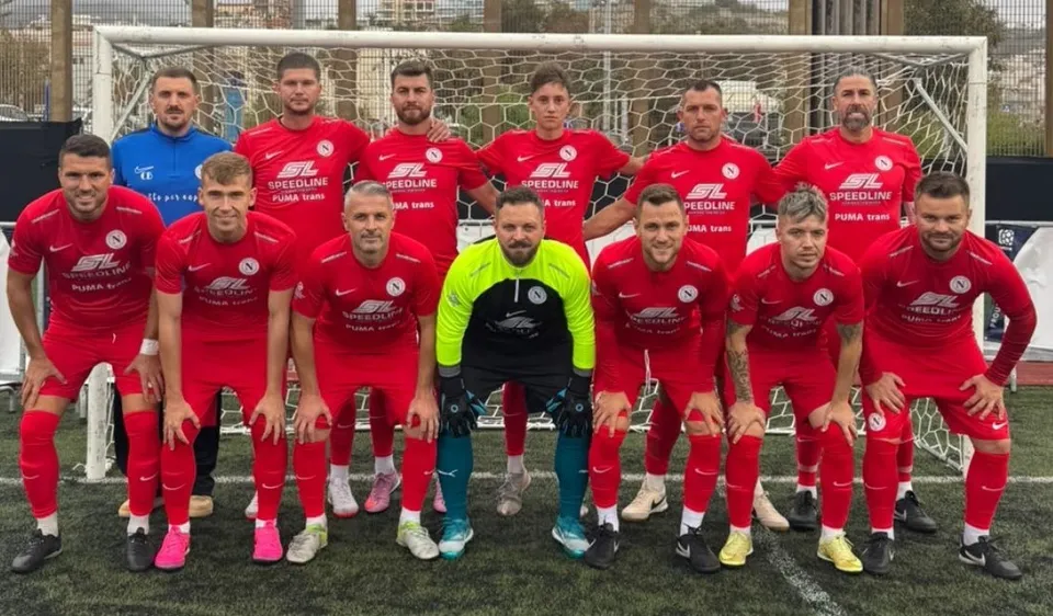O echipă de fotbaliști în echipamente roșii și un jucător în echipament galben stau în poziție de pregătire în fața unui por.
