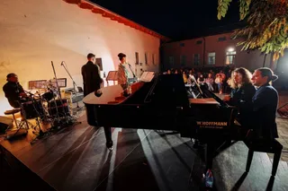 O scenă de concert în aer liber noaptea, cu un pian de concert negru, un baterist la stânga, cântăreți pe scenă și un public așezat în fundal.