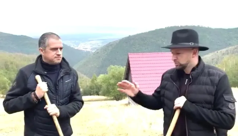 Un bărbat cu pălărie neagră și o vestă neagră gesticulează către un alt bărbat care ține o lopată în fața unor dealuri verzi.