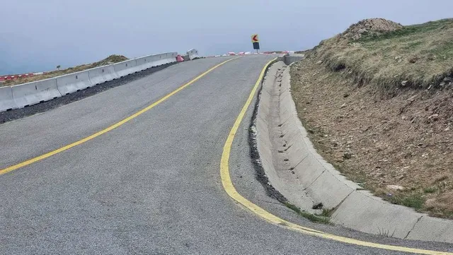 O porțiune de drum asfaltat cu linii galbene continue curbează spre dreapta, fiind delimitată de un gard de beton și un talcu de pământ cu iarbă.