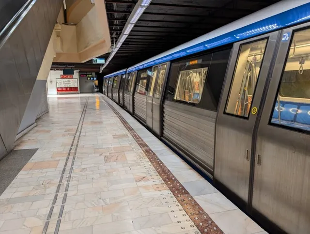 Un tren de metrou gri cu ferestre mari stă oprit pe un peron de stație cu podea din plăci de marmură deschisă.