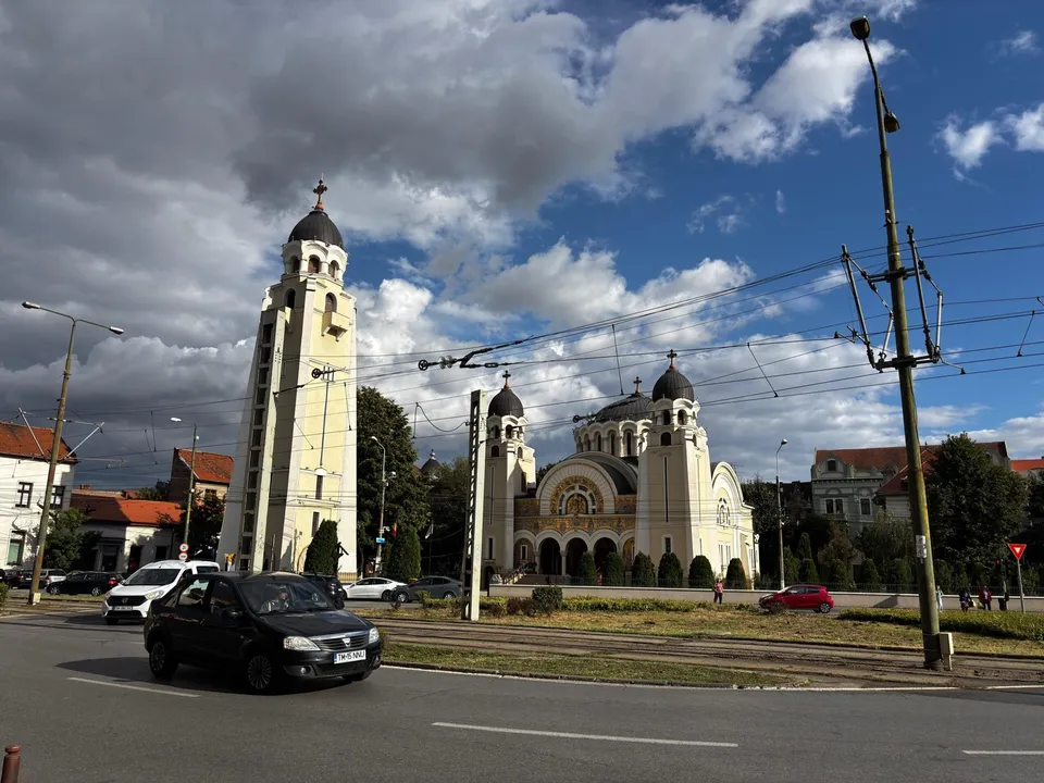 O biserică cu turnuri înalte și cupole se află lângă o stradă cu mașini în prim-plan și linii electrice suspendate sub un cer noros.