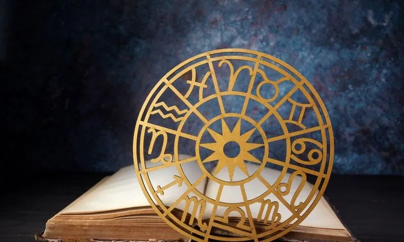 Un obiect circular din lemn cu simboluri ale zodiacului este așezat deasupra unei cărți deschise pe o suprafață întunecată.