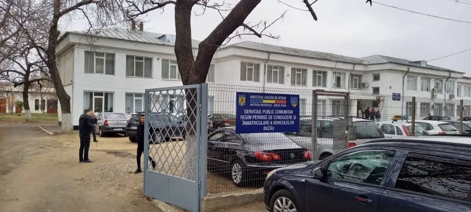 O clădire albă cu ferestre rectangulare este vizibilă în spatele unui gard metalic și a unor mașini parcate pe marginea unui drum.