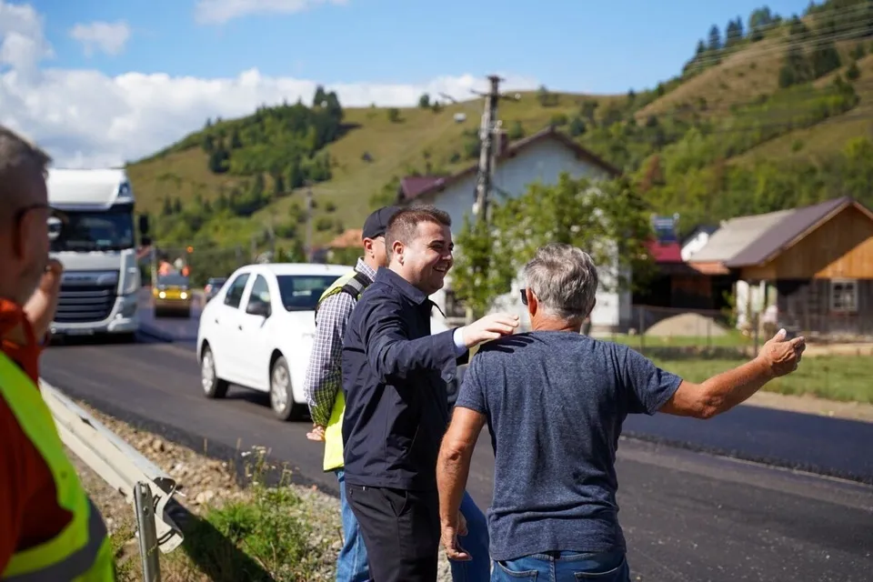 Un grup de bărbați stă pe marginea unui drum asfaltat lângă o mașină albă, sub un cer cu nori și dealuri verzi.