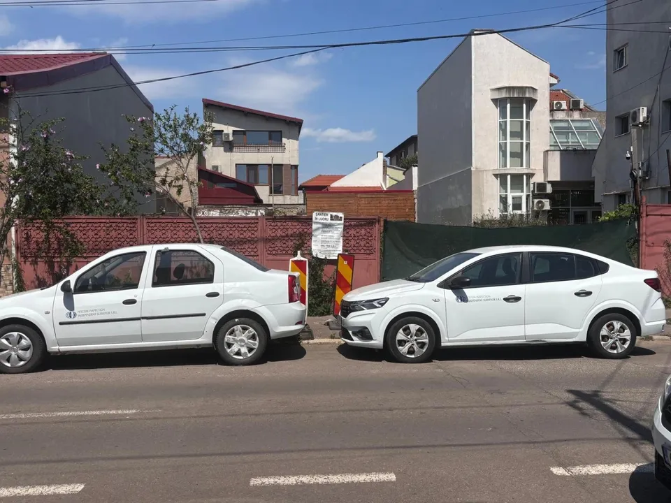 Două mașini albe sunt parcate pe marginea unui drum asfaltat în fața unor clădiri și garduri.