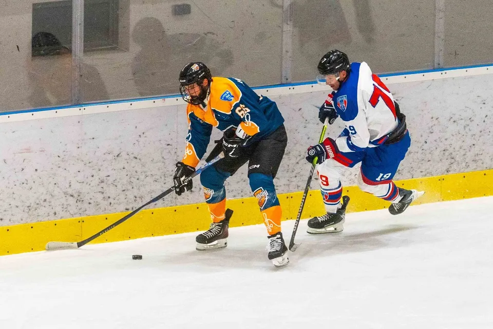 Un jucător de hochei cu echipament albastru și portocaliu conduce un puck pe gheață, fiind urmărit de un adversar în echipament alb, albastru și roșu.