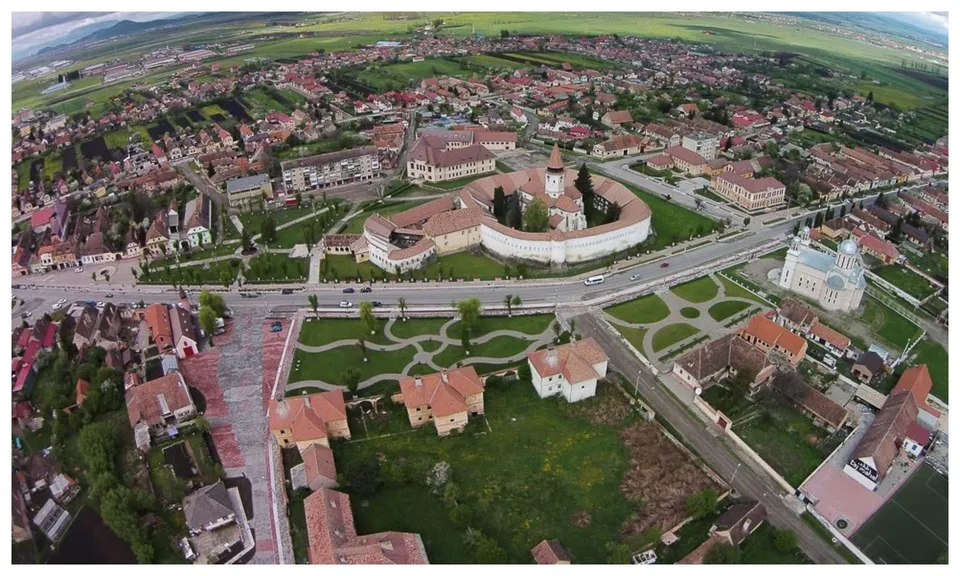 O vedere aeriană prezintă o localitate cu case cu acoperișuri colorate, o clădire circulară cu ziduri albe și câteva biserici în jur.