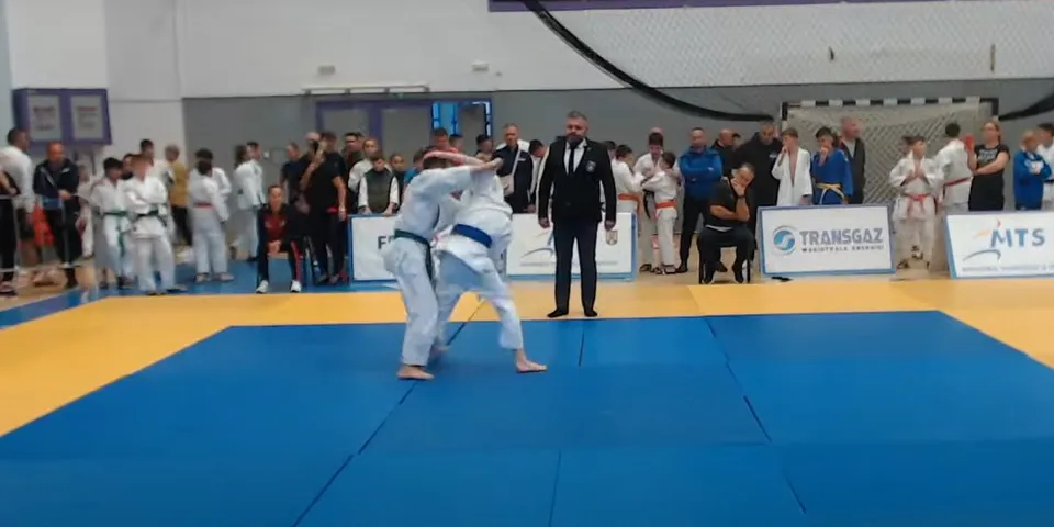 Doi judoka se luptă pe un tatami albastru și galben, în timp ce un arbitru și spectatori privesc de pe margine, lângă panouri publicitare.