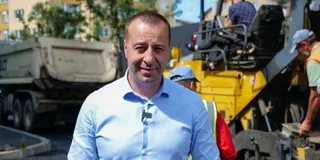 Un bărbat cu cămașă albastră stă în prim-plan, având în fundal un camion și utilaje de construcții galbene.