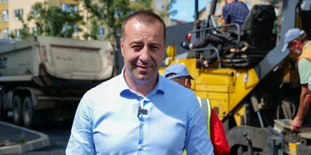 Un bărbat cu cămașă albastră stă în prim-plan, având în fundal un camion și utilaje de construcții galbene.
