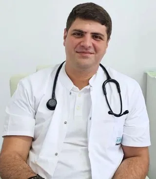 Un bărbat cu părul scurt și întunecat poartă un halat alb peste un tricou alb și are un stetoscoap negru în jurul gâtului.