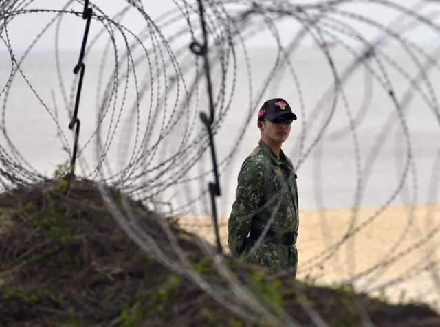 Un bărbat îmbrăcat în uniformă militară de tip camuflaj stă pe o plajă lângă un munte de pământ, fiind parțial vizibil prin sârmă ghimpată.