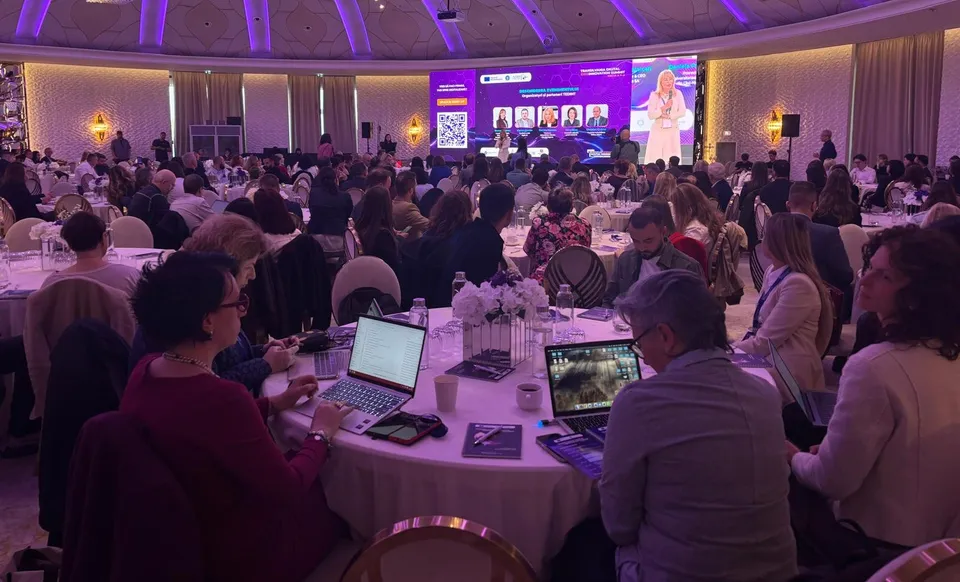 Oamenii stau la mese rotunde cu laptopuri într-o sală de conferințe mare cu un ecran digital luminos în fundal.