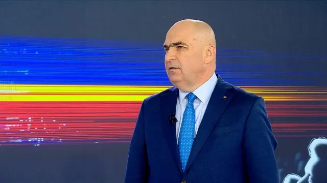 Un bărbat în costum albastru cu cravată albastră este înfățișat dintr-un unghi lateral în fața unui ecran cu drapelele României.
