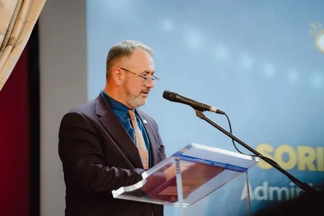 Un bărbat cu ochelari și barbă stă în spatele unui podium transparent și vorbește la un microfon.
