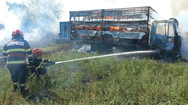 Două persoane cu echipament de protecție și căști roșii direcționează un jet de apă către un autocamion care arde într-un câmp cu vegetație.
