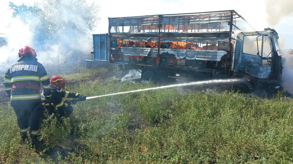 Două persoane cu echipament de protecție și căști roșii direcționează un jet de apă către un autocamion care arde într-un câmp cu vegetație.