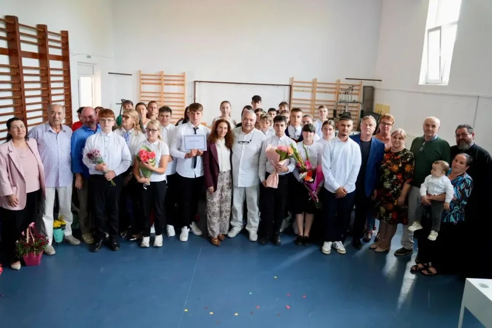 Un grup de adulți și copii stă aliniat în sală, unii ținând buchete și o diplomă.
