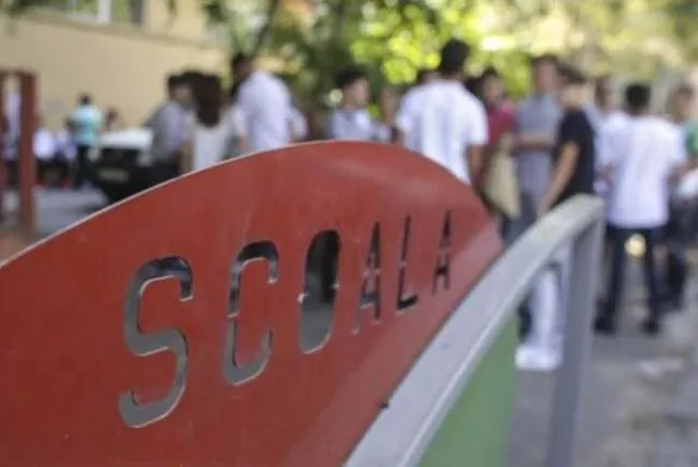 Un semn roșu cu textul metalic sculptat este în prim-plan, în timp ce un grup de persoane stă în fața unei clădiri în fundalul neclar.