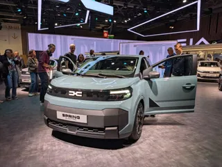 Un autoturism de culoare albastru deschis cu portierele deschise este expus într-un spațiu de expoziție cu oameni în fundal.
