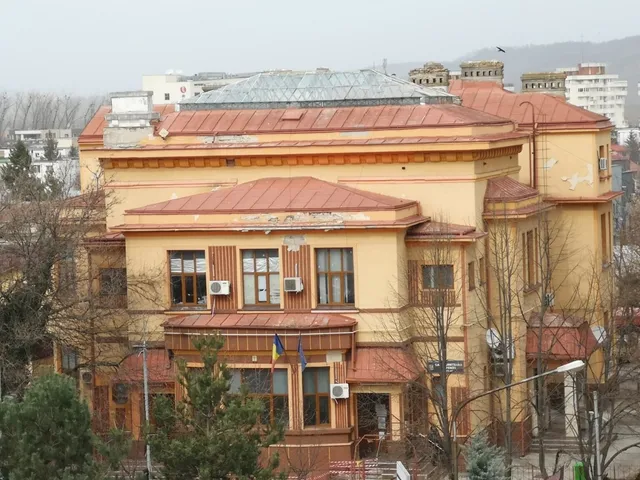 O clădire de culoare galben cu acoperiș de culoare maro și ferestre cu rame maro este înconjurată de copaci fără frunze sub un cer gri.