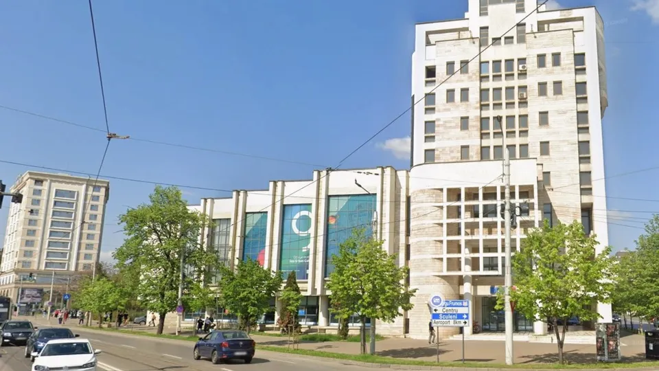 Imaginea prezintă o clădire înaltă, albă, cu nouă etaje, înconjurată de copaci și o stradă circulată, sub un cer senin.