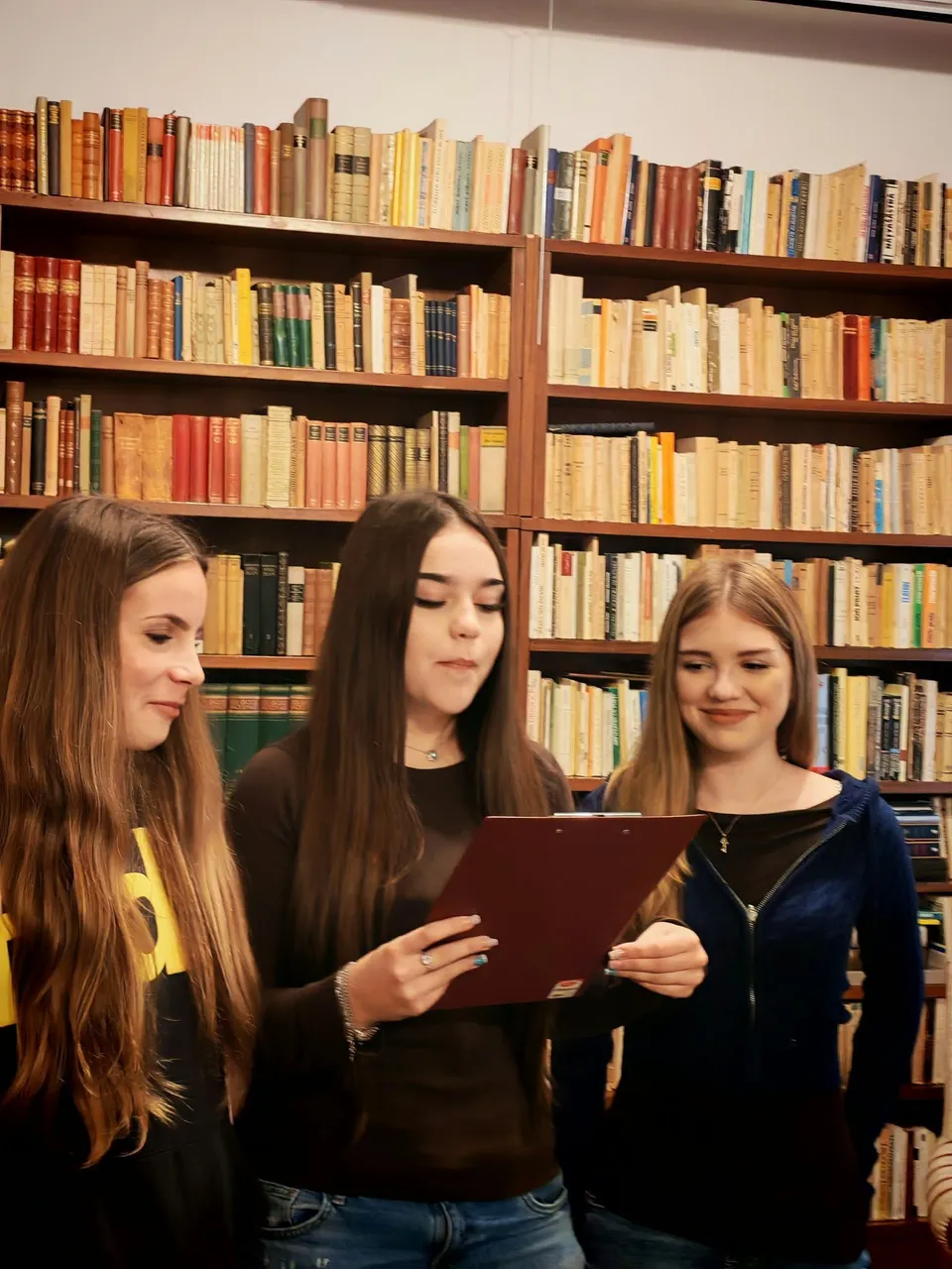 Trei fete tinere, în fața unor rafturi de carte pline, se uită atent la o tabletă pe care o ține fata din mijloc.