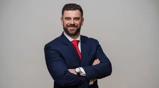 Un bărbat cu barbă, purtând un costum albastru, cămașă albă și cravată roșie, stă cu brațele încrucișate în fața unui fundal gri neutru.